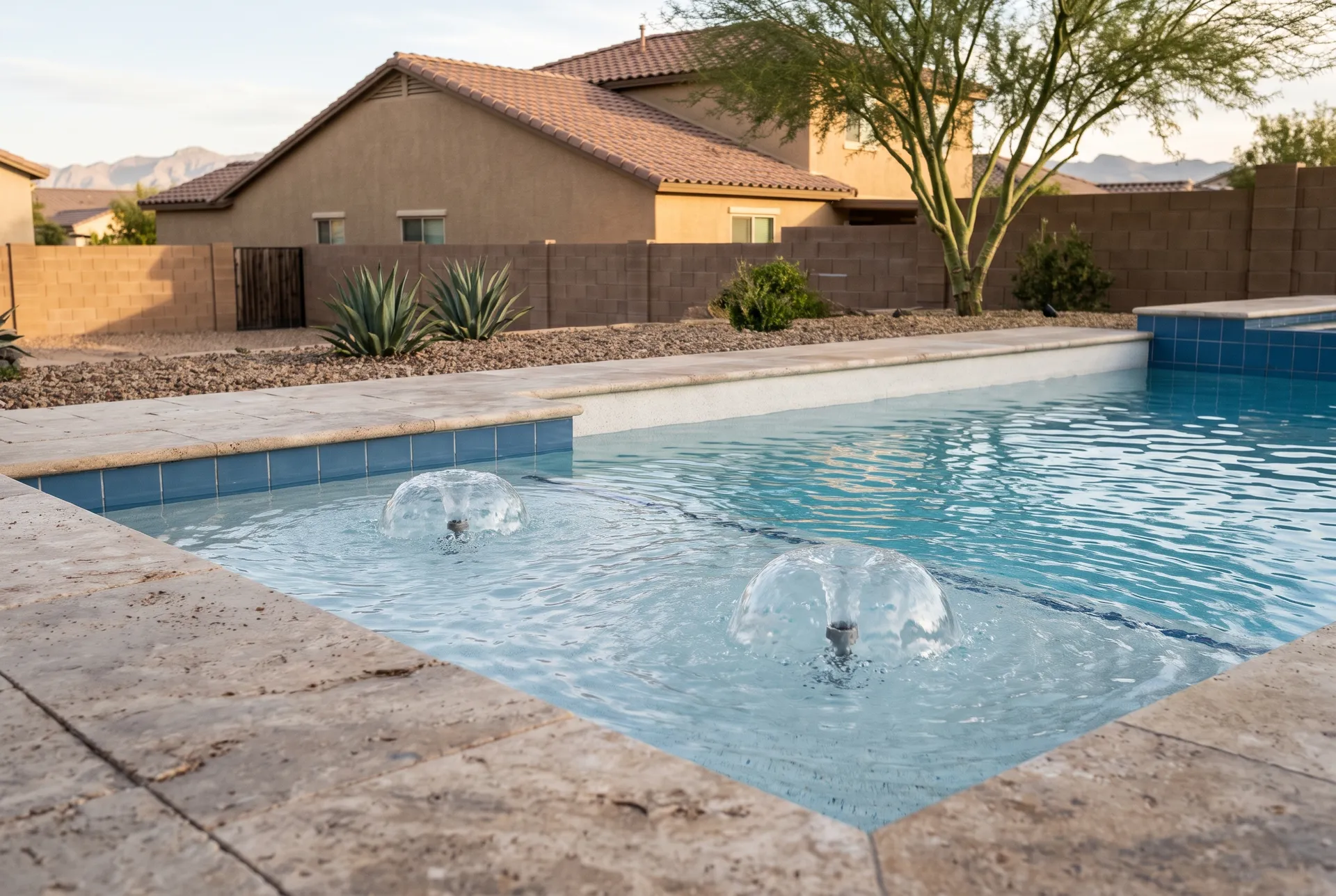 Two small bubbler water features on a shallow tanning ledge of a backyard pool, gentle domes of water, clear blue water, travertine deck