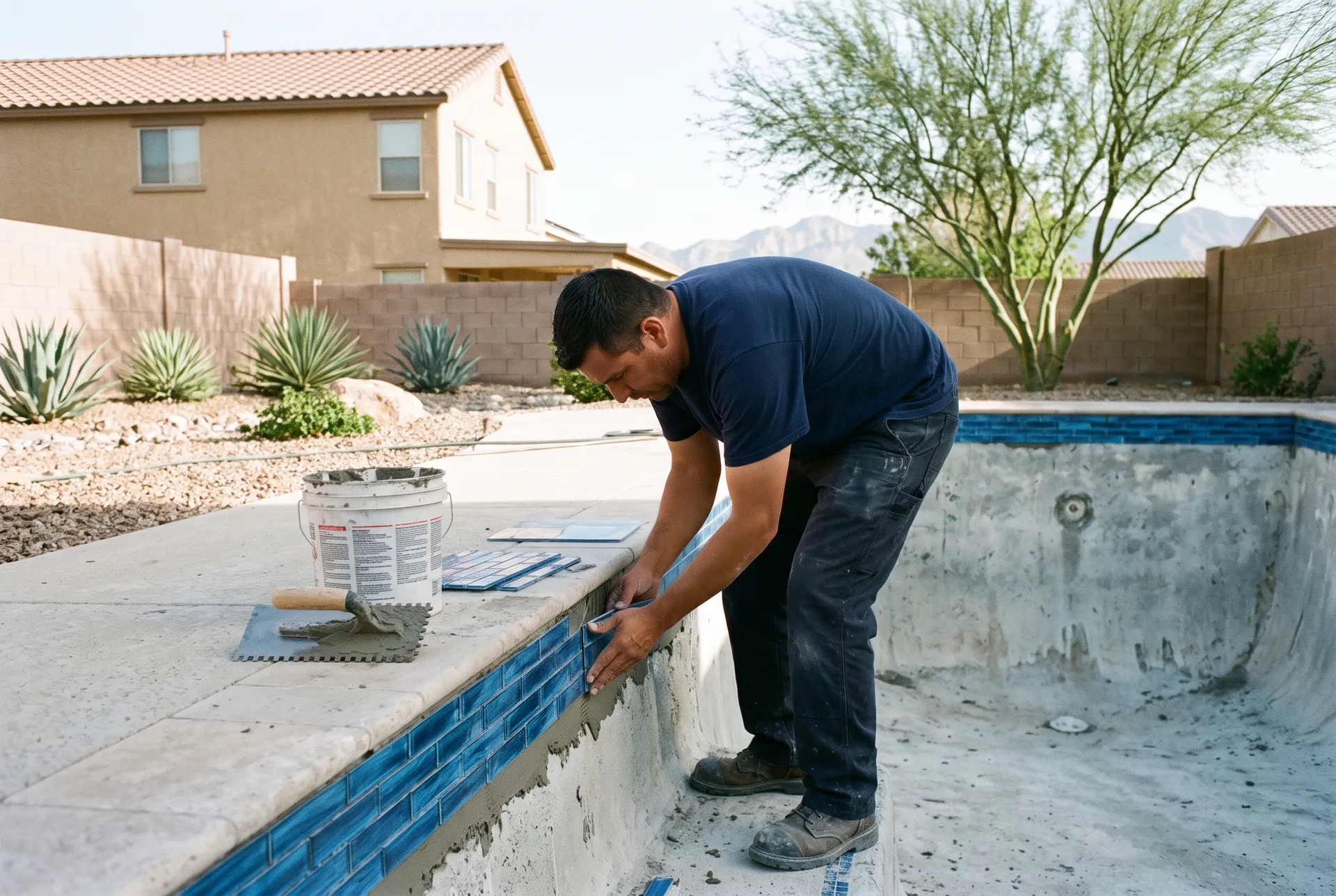 A tile setter in a navy shirt pressing new blue glass waterline tiles onto the upper wall of a drained pool