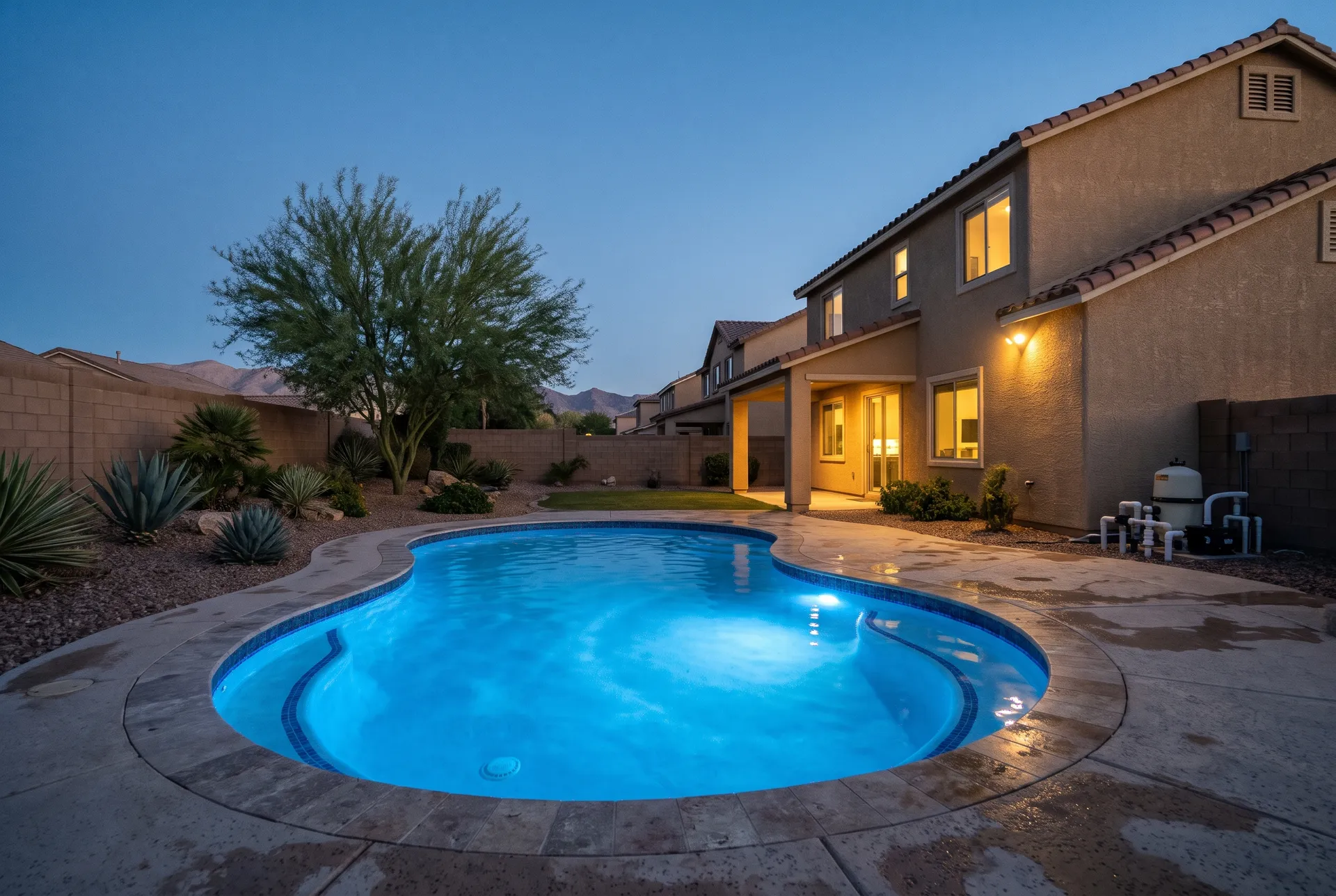 A backyard pool at dusk with an underwater LED light glowing blue, the water lit from within, the home lights on, mountains silhouetted
